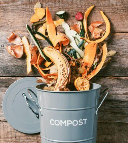 Peeled vegetables on chopping board, white compost bin on wooden background. Top view of kitchen